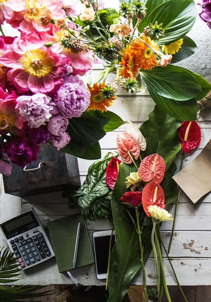 Various flowers, including pink peonies and orange daisies, arranged on a rustic wooden table with a calculator, notebook, pen, smartphone, and envelopes.