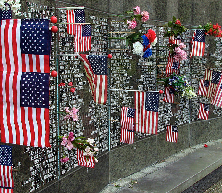 American flags and bouquets of flowers adorn a black granite memorial wall engraved with numerous names, honoring fallen soldiers. This solemn tribute creates a poignant atmosphere of remembrance and respect.