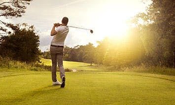 A golfer, mid-swing, stands on a lush green course under a bright setting sun, surrounded by trees and open fields.