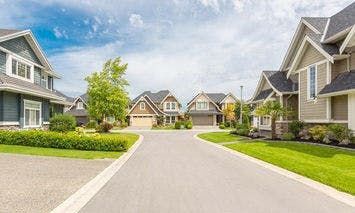 Modern suburban houses line both sides of a curved, paved street, with landscaped lawns and trees, under a partly cloudy sky. The neighborhood appears clean and well-maintained.