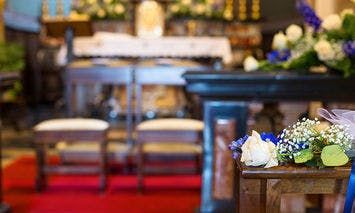 Flowers rest on a wooden pew in a decorated church, with an altar adorned with more flowers and a red carpet leading up to it in the background.