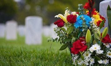 A bouquet of vibrant flowers rests against a white tombstone in a cemetery, with additional blurred tombstones visible in the background.