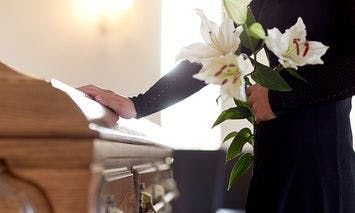 A person in black attire stands beside a wooden casket, holding white lilies in one hand, in a softly lit room.