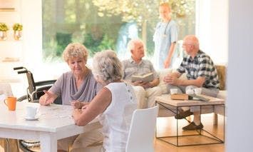 Seniors are sitting at a table and conversing in a brightly lit, cozy room. Another group is chatting on a couch with a caregiver standing nearby in the background.