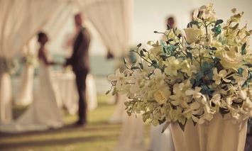 A bouquet of white and blue flowers rests in the foreground; in the blurred background, a couple stands facing each other under a white draped canopy on a grassy area.